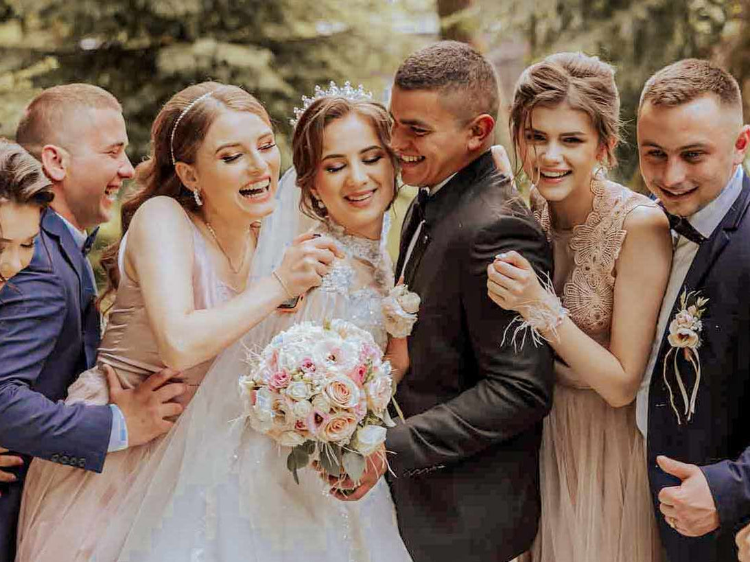 A joyful wedding party poses together outdoors, with the bride and groom surrounded by smiling bridesmaids in blush dresses and groomsmen in dark suits, celebrating in a close, affectionate group hug.